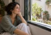 Mujer escuchando el entorno junto a una ventana abierta.