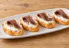 Pintxo de anchoa y crema de queso Filetes de anchoa plegados sobre queso crema blanco y rebanadas de pan.