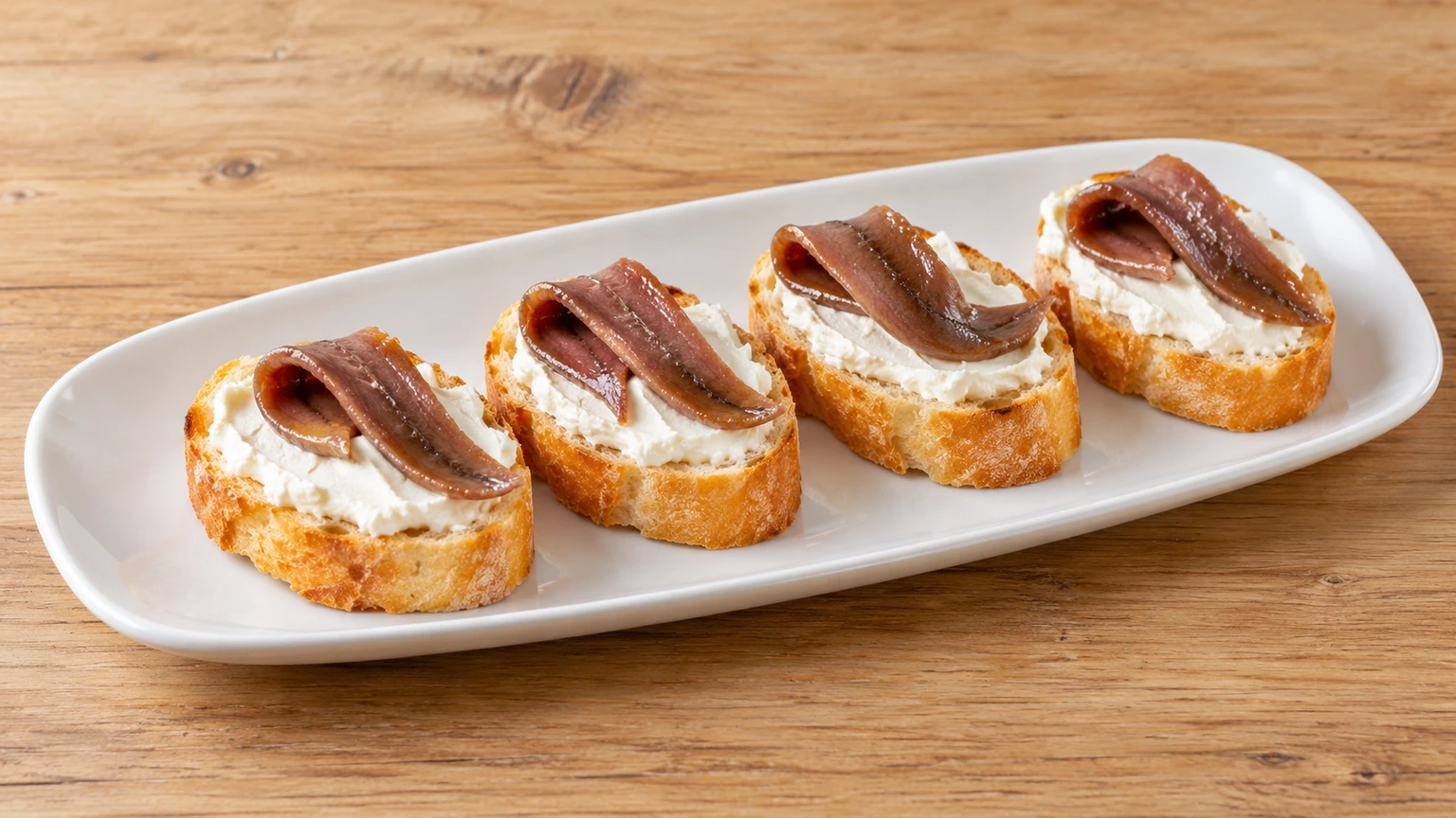 Filetes de anchoa plegados sobre queso crema blanco y rebanadas de pan.