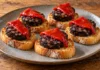 Pintxo de morcilla y piquillo Rodajas doradas de morcilla de arroz sobre pan con tiras rojas de piquillo.