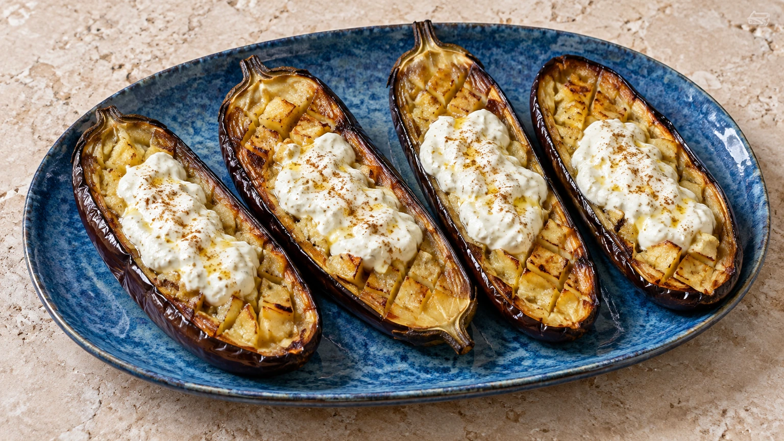 Mitades de berenjena asada con cortes en rombos y salsa blanca de yogur sobre la pulpa dorada.