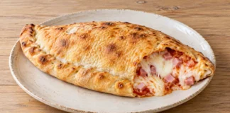 Calzone de jamón y queso Calzone dorado abierto por un extremo con jamón cocido, mozzarella fundida y tomate visibles.
