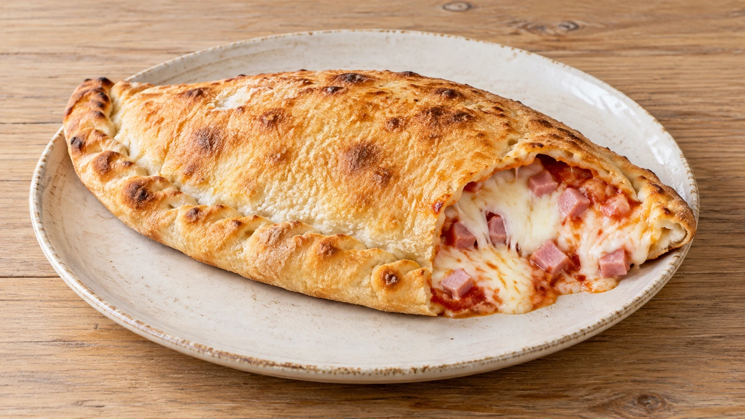 Calzone dorado abierto por un extremo con jamón cocido, mozzarella fundida y tomate visibles.